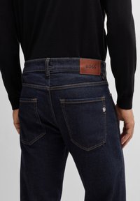 Dark blue denim jeans with a brown leather label featuring "BOSS" on the waistband. Stitching lines are visible on back pockets.