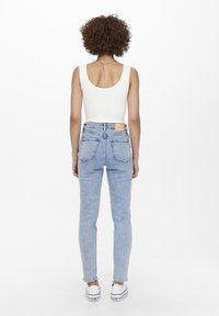 Persona con cabello rizado, usando una camiseta sin mangas blanca, jeans de tiro alto azul claro y zapatillas blancas, de pie de espaldas sobre un fondo blanco.