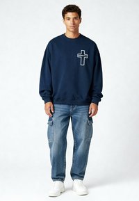Jeune homme portant un sweat-shirt marine avec le logo croisé "Karma Kills", un jean cargo bleu et des baskets blanches, debout devant un fond uni.