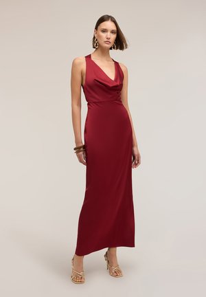 Frau trägt ein ärmelloses, knöchellanges Satinkleid in tiefem Rot mit drapiertem Ausschnitt, goldenen Ohrringen, Armbändern und beigen Sandalen mit Absatz.