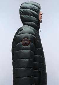 Donkergroene gewatteerde jas met een capuchon, horizontaal gestikte patroon, ritszakken en een ronde patch op de bovenarm.