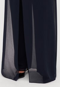 Tamsiai mėlyna permatoma maksi suknelė su ilgu iškirpimu. Audinys atrodo lengvas ir elegantiškai krenta, atidengdamas tvirtą pamušalą ir avalynę.