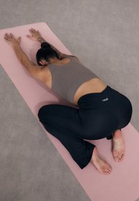 Black yoga pants and a brown tank top worn by a person in a forward stretch position on a pink yoga mat, with a light gray surface below.