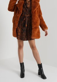 Brązowy płaszcz z sztucznego futra z dużym kołnierzem, noszony na wzorzystej sukience w ciepłych tonach, zestawiony z czarnymi botkami na kostkę o fakturze krokodyla.