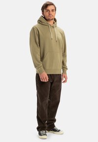 Olive green hooded sweatshirt with drawstrings, front kangaroo pocket, and ribbed cuffs, paired with brown corduroy pants and black sneakers.