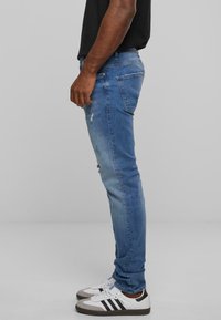 Férfi kék farmernadrág, slim fit szabással, kopott részletekkel. Anyaga pamut, enyhe elasztikus hatással. Fehér tornacipővel és fekete inggel viselve.