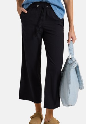 Personne portant un pantalon noir ample et des chaussures slip-on couleur camel, tenant un sac fourre-tout bleu clair dans une main.