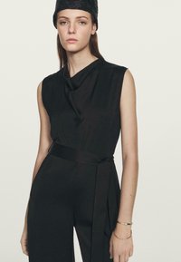 Zwarte mouwloze jumpsuit met een gedrapeerde halslijn, een ceintuur in de taille en een zachte textuur. Gemaakt in combinatie met een gestructureerde zwarte hoed en minimalistische gouden armbanden.