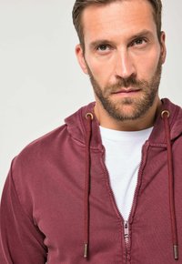 Sudadera con capucha de cremallera color burdeos con cordones, con una textura suave y camiseta blanca debajo. Detalle de cremallera visible en la parte delantera.