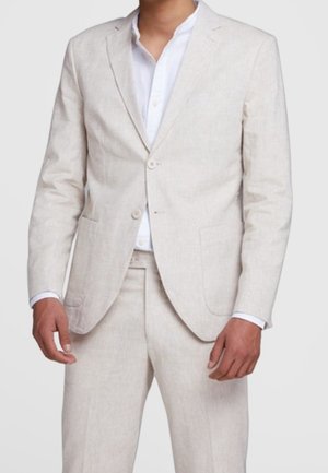 Homme portant un costume beige clair avec une chemise blanche, veste boutonnée et pantalon assorti, debout devant un fond uni.