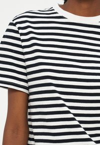 Chemise à manches courtes rayée horizontalement en noir et blanc portée par une personne à la peau foncée sur un fond uni.