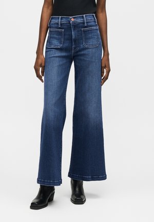 Γυναίκα που φοράει τζιν με ψηλή μέση και φαρδιά πόδια σε σκούρο μπλε χρώμα, με μπροστινές τσέπες και μαύρα μποτάκια αστραγάλου, στέκεται μπροστά σε απλό φόντο.