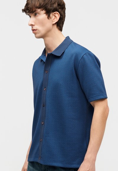 Jeune homme portant une chemise bleue texturée à manches courtes avec col, debout de profil devant un fond uni.