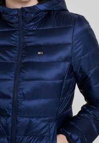 Person bærende en marineblå quiltet jakke med hætte og logo-plekt på brystet, med hånden stukket ind i sidelommen.