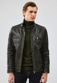 Veste en cuir noir avec une finition brillante, deux poches zippées sur la poitrine et des coutures contrastées. Stylée avec un col roulé vert en dessous.