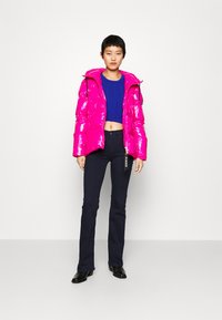 Slik, lys rosa dunjakke med hette, glidelås foran og justerbare snor, kombinert med en blå kortermet genser og mørke jeans.