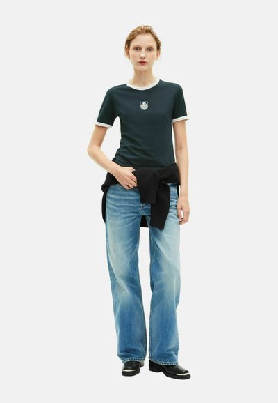 Dunkelgrünes T-Shirt mit weißem geripptem Kragen und Logo, kombiniert mit hellblauen weit geschnittenen Jeans und schwarzen Stiefeletten. Schwarzer Pullover am Bund gebunden.