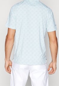 Košile s krátkým rukávem zobrazená z boku, s světle modrým geometrickým vzorem, hladkým povrchem tkaniny a bočními rozparky.