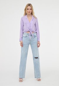 Blusa lunga di colore viola chiaro annodata in vita, abbinata a jeans a gamba larga blu chiaro con un strappo al ginocchio sinistro. Sandali bianchi.