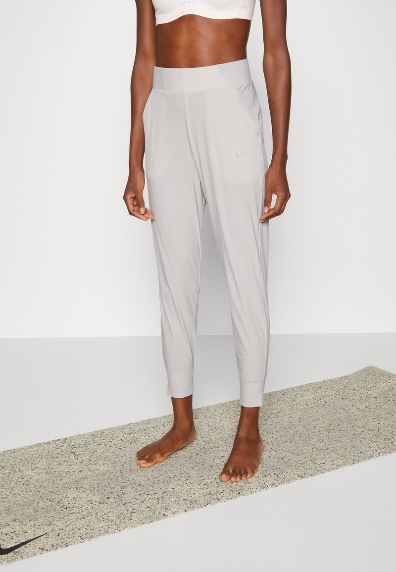 Pantalones deportivos de color gris claro con un corte ajustado, puños cónicos, dos bolsillos laterales y un pequeño logotipo de Nike en negro en el lado izquierdo. Textura suave y lisa.