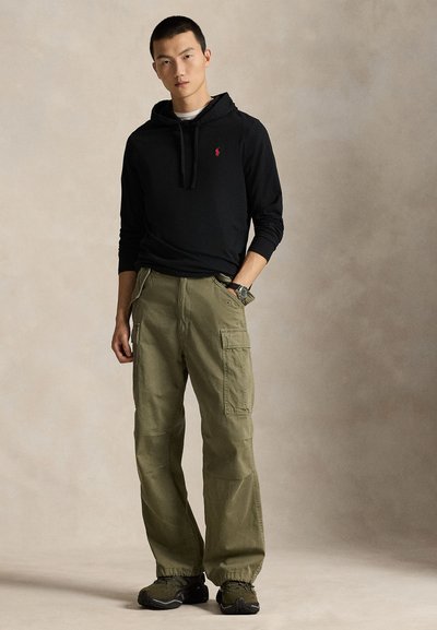 Moški, ki nosi črno kapucar s rdečim logotipom, olive cargo hlače in pohodniške čevlje, stoji z eno roko v žepu pred bež ozadjem.