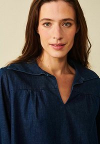 Mörk denimtop med bred krage, plisserade detaljer under bröstet och V-ringning. Slät struktur och subtil lyster är tydlig.