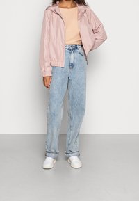 Giacca rosa chiaro con zip e polsini elasticizzati, abbinata a jeans blu chiaro dal taglio morbido e sneakers bianche. Tessuto liscio e design informale.