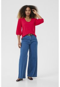 Roter Pullover mit kurzen, puffärmeligen Ärmeln, kombiniert mit hochgeschnittenen blauen weit geschnittenen Jeans und einem braunen Gürtel. Dunkelbraune Schuhe runden das Outfit ab.
