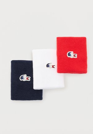 Tre polsini di cotone piegati nei colori blu navy, bianco e rosso, ciascuno con una piccola toppa con il logo del coccodrillo nei colori blu, bianco e rosso.