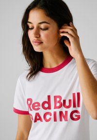 Tricou alb din bumbac cu guler și manșete roșii. Prezentă un text îndrăzneț roșu "Red Bull RACING" pe partea din față.