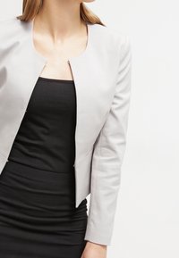 Veste grise texturée avec un col rond, une fermeture éclair à l'avant et des manches longues, portée par-dessus une robe noire ajustée.