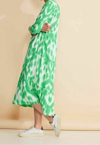 Vihreä kuviollinen mekko, jossa on virtaava, kerroksellinen muotoilu ja pitkät hihat, yhdistettynä valkoisiin lenkkareihin. Sileä kangas ikat-kuviolla.