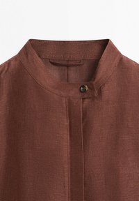 Col de chemise en tissu marron texturé avec un seul bouton noir de fixation sur un col mandarin, présenté sur un fond blanc.