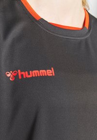 Camiseta deportiva negra con cuello redondo y un acento naranja. El logotipo de la marca "hummel" se muestra prominentemente en rojo en el frente.