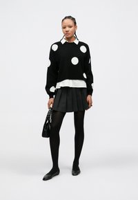 Μαύρο πουλόβερ με λευκές πουά, φορεμένο πάνω από λευκό πουκάμισο. Μαύρη μίνι φούστα με πιέτες, μαύρα καλσόν και μαύρα επίπεδα παπούτσια. Μικρή τσάντα χειρός.