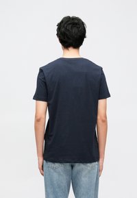 Tumši zila īsroku kokvilnas T-krekls ar apaļu kaklu un brīva piegriezuma siluetu, redzams no aizmugures. Nav redzamu rakstu vai logotipu.