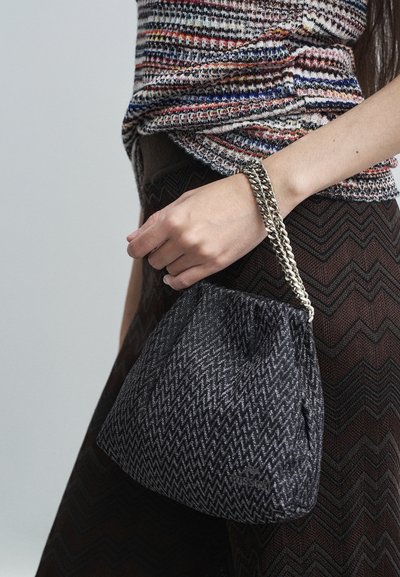 Woman holding a small black and gray zigzag-patterned handbag with a silver chain handle, wearing a multicolored textured top and dark zigzag skirt.