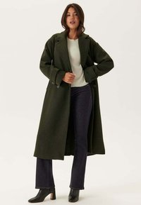 Cappotto in misto lana verde oliva con design doppiopetto, ampi rever e due bottoni, abbinato a un maglione leggero e jeans scuri.