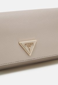 Lichtbeige portemonnee met een gestructureerd oppervlak, voorzien van een gouden driehoekig embleem met "GUESS USA." Gestikte details langs de randen.