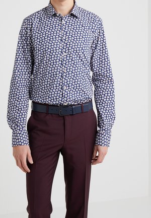 Uomo che indossa una camicia a maniche lunghe blu con motivo floreale infilata in pantaloni color bordeaux con cintura blu scuro, in piedi davanti a uno sfondo semplice.