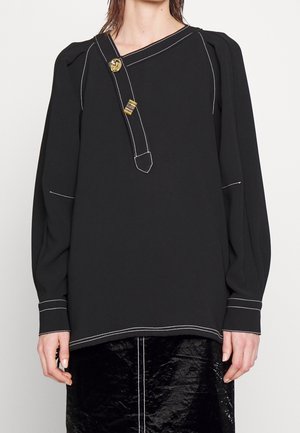 Czarna bluzka z długim rękawem z białym przeszyciem, asymetrycznym dekoltem, dwoma złotymi ozdobnymi klipsami, noszona z czarną błyszczącą spódnicą.