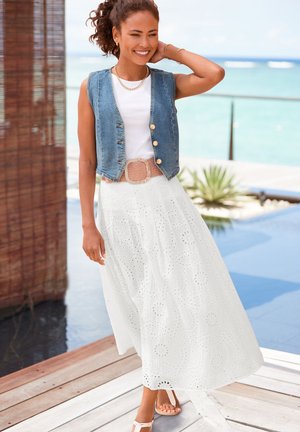 Ärmellose Jeansweste, weißer Maxirock mit Lochstickerei, hellbrauner geflochtener Gürtel, helle Sandalen. Modell steht in einer hellen Außenumgebung.
