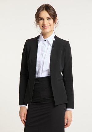 Jonge vrouw glimlacht, draagt een zwarte blazer, wit hemd met knopen en een zwarte rok, staand tegen een effen lichte achtergrond.