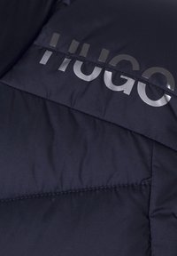 Zbliżenie na ciemnoniebieską pikowaną kurtkę z przeszyciami oraz częściowo widocznym błyszczącym logo marki "HUGO" na powierzchni materiału.