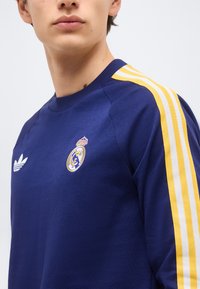 Persona con sudadera azul marino con franjas amarillas y blancas en los hombros y logotipos deportivos en el pecho.