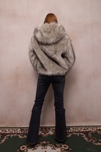 Person with long hair wearing a large gray fur coat and dark flared jeans standing on a patterned carpet against a plain wall.