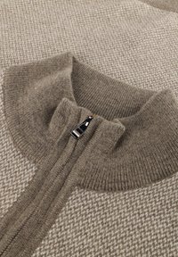 Nahaufnahme eines beigen und braunen Strickpullovers mit einem gerippten hohen Kragen und einem silbernen Reißverschluss-Zieher.