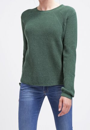 Kobieta ubrana w dopasowany, teksturowany ciemnozielony sweter z dzianiny z długimi rękawami oraz niebieskie dżinsy, stojąca na tle jednolitego, jasnego tła.