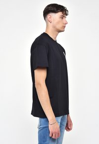 Musta lyhythihainen puuvillapaita, jossa on pyöreä kaula-aukko ja pieni valkoinen logo rinnassa. Leikkaus on rento ja suora.