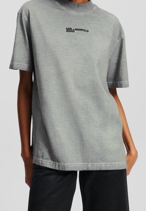 Personne portant un T-shirt oversize gris clair avec le logo « KARL LAGERFELD JEANS » centré sur la poitrine, associé à un pantalon noir.